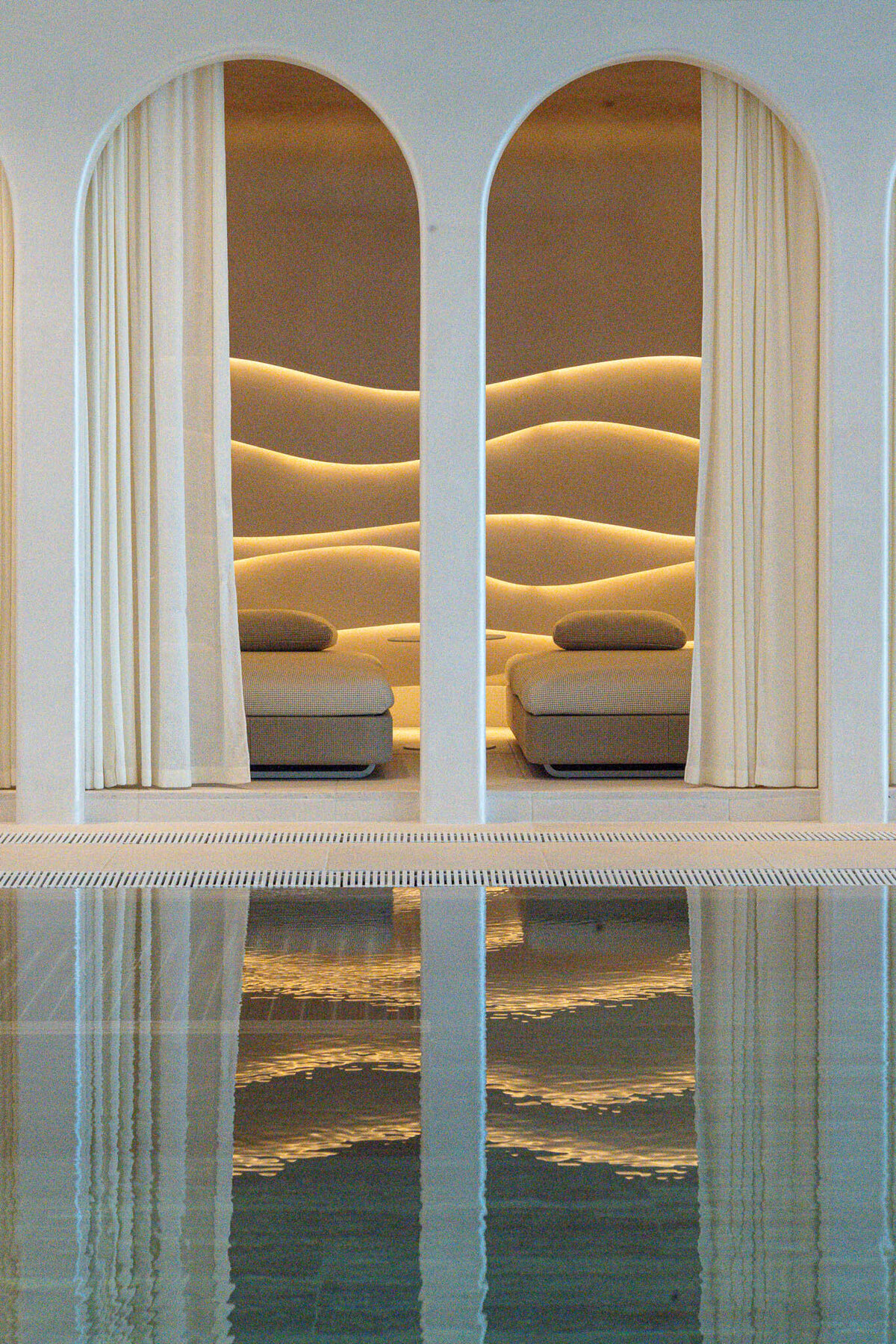 Luxurious spa interior with arched doorways and an illuminated wavy wall design behind spa beds. Sheer curtains partially cover the openings. Calm water in a pool reflects the serene setting, enhancing the tranquil ambiance.