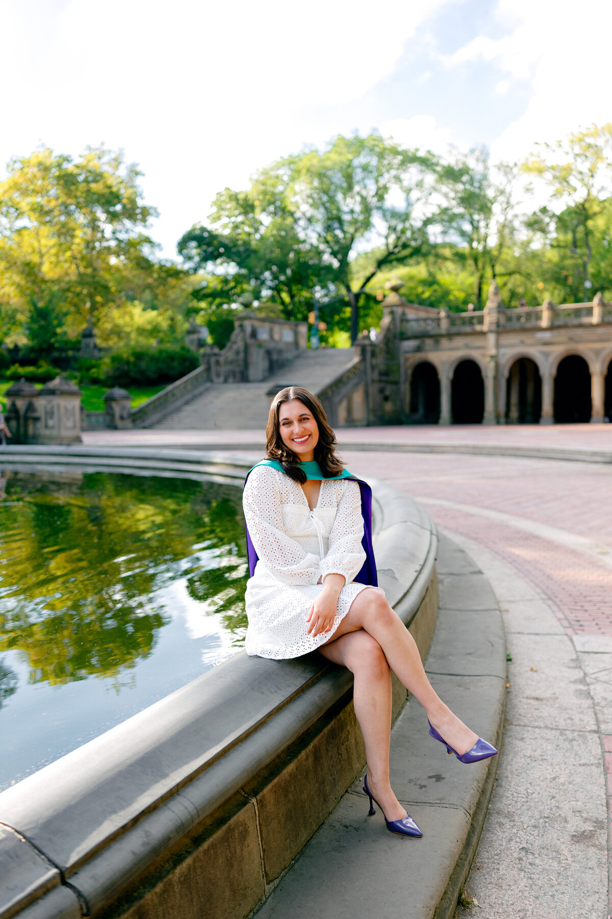 NYU graduate smiling at the camera in white dress and purple heels during sunrise at Central Park.