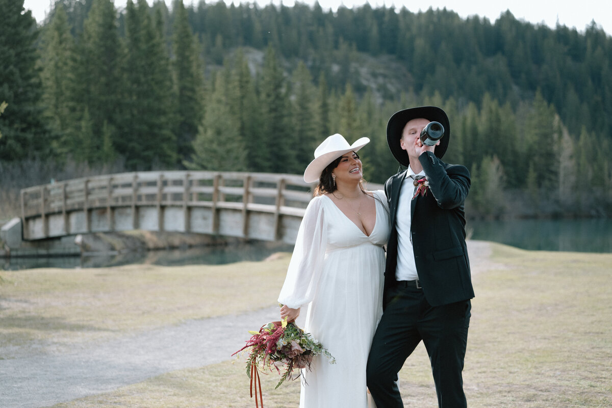 Newlyweds walk near a wooden bridge with mountains and forest surrounding them in Banff popping a bottle of champagne.