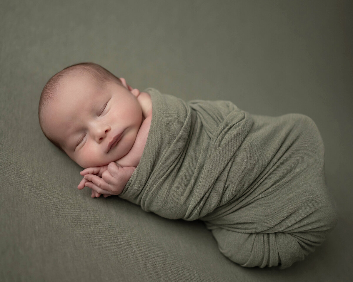 A newborn baby sleeps in a tight green swaddle in a studio of an Iowa Newborn Photographer
