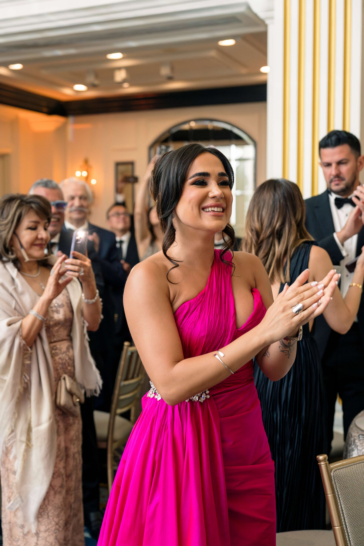 A woman in a vibrant pink dress claps her hands, smiling in a room filled with well-dressed people. Others around her appear to be socializing and enjoying a formal event. The setting has elegant decor with mirrors and warm lighting.