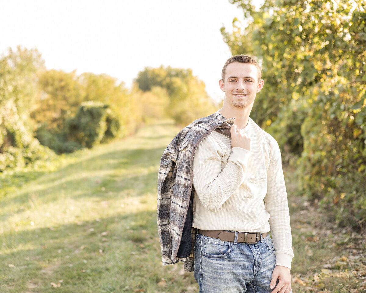 high school senior male, standing in grass with trees behind him, with a flannel jacket over his shoulder, wearing a light tan, long sleeve shirt, blue jeans, and a belt, looking slightly off camera smiling