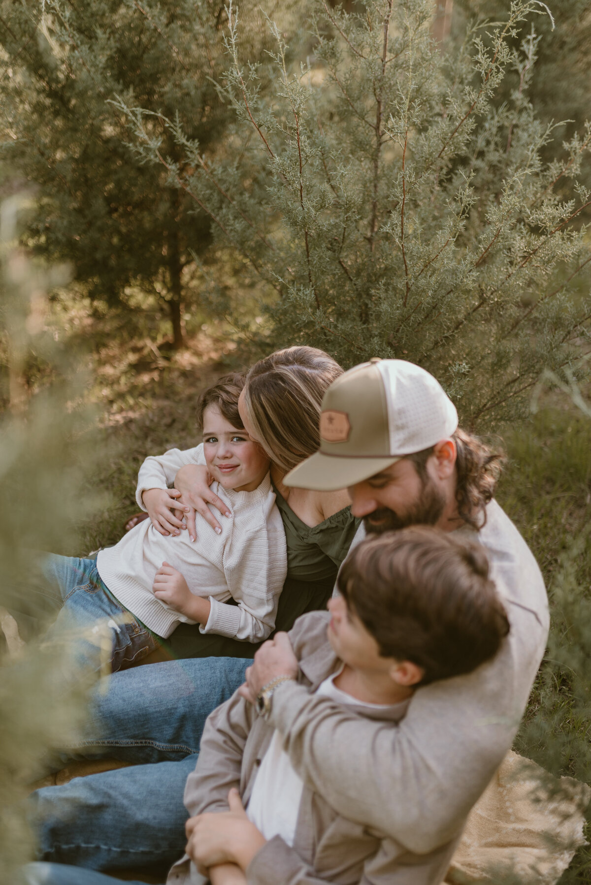 a family of 4 sitting on a blanket at a christmas tree farm, tickling each other and laughing