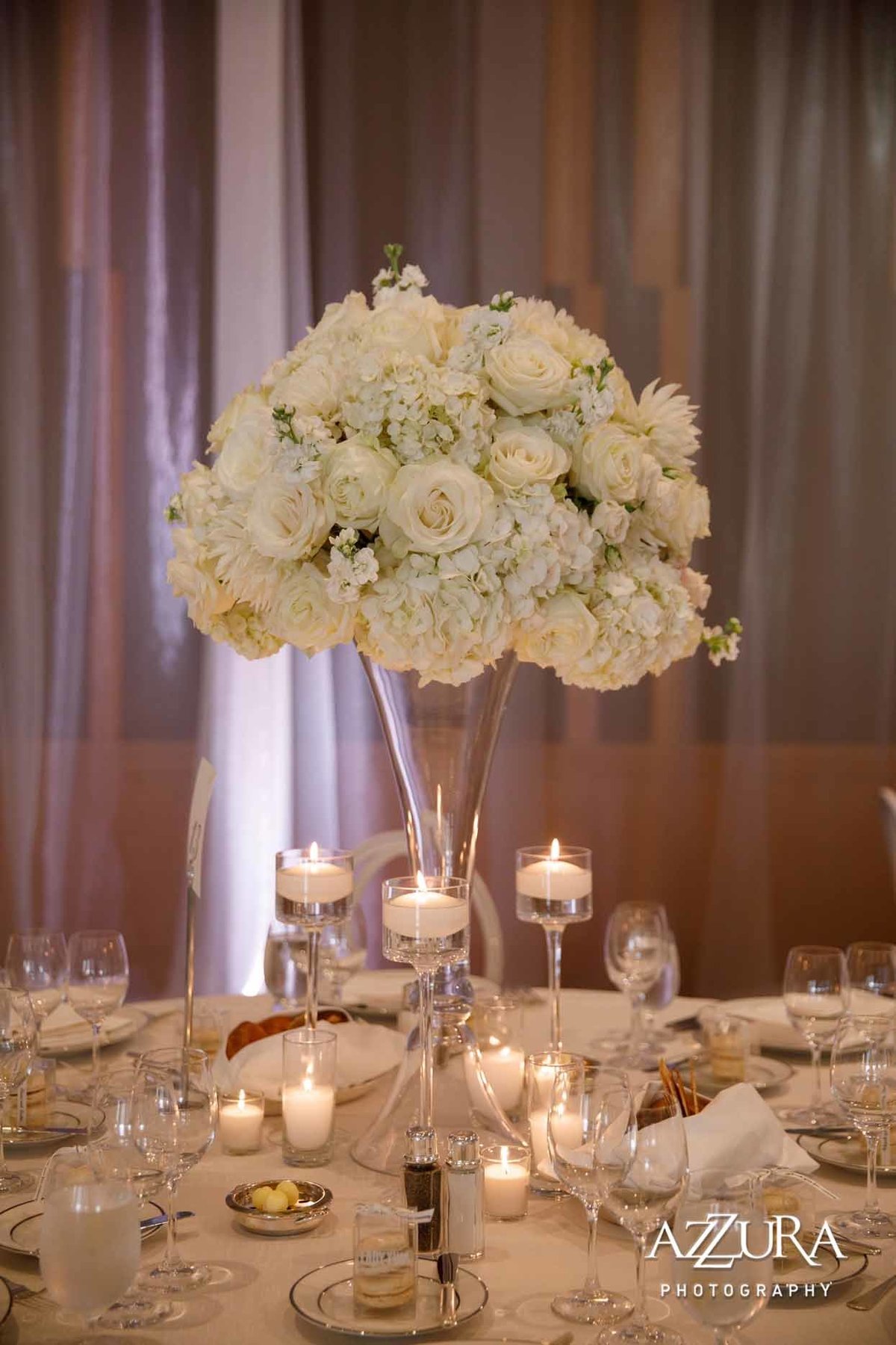 large white centerpiece of all white roses, white hydrangea, white stock on tall crystal vase with glass candle holders on white linen