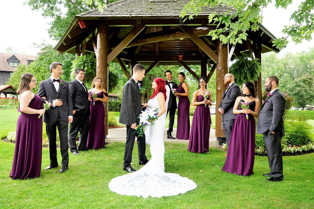 The bride with fiery red hair gazes lovingly at the groom while surrounded by the bridal party. The scene captures a moment of intimate connection and joy amidst a festive group, showcasing the bride's unique hair color and the supportive presence of the bridal party.