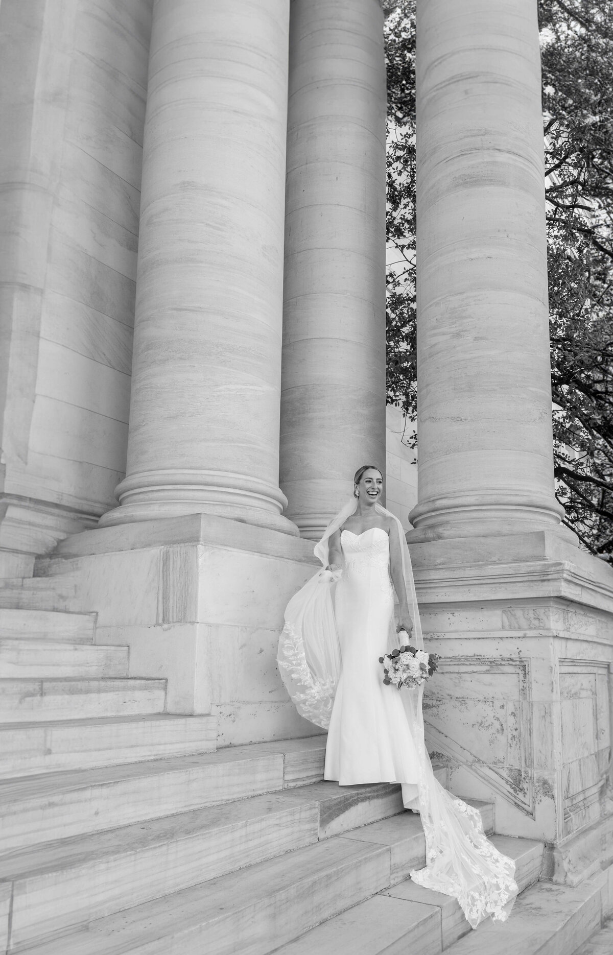 A bride in a white wedding gown stands on the steps of a grand building with large columns. She holds a bouquet and her long veil drapes down the steps. Black and white photo.