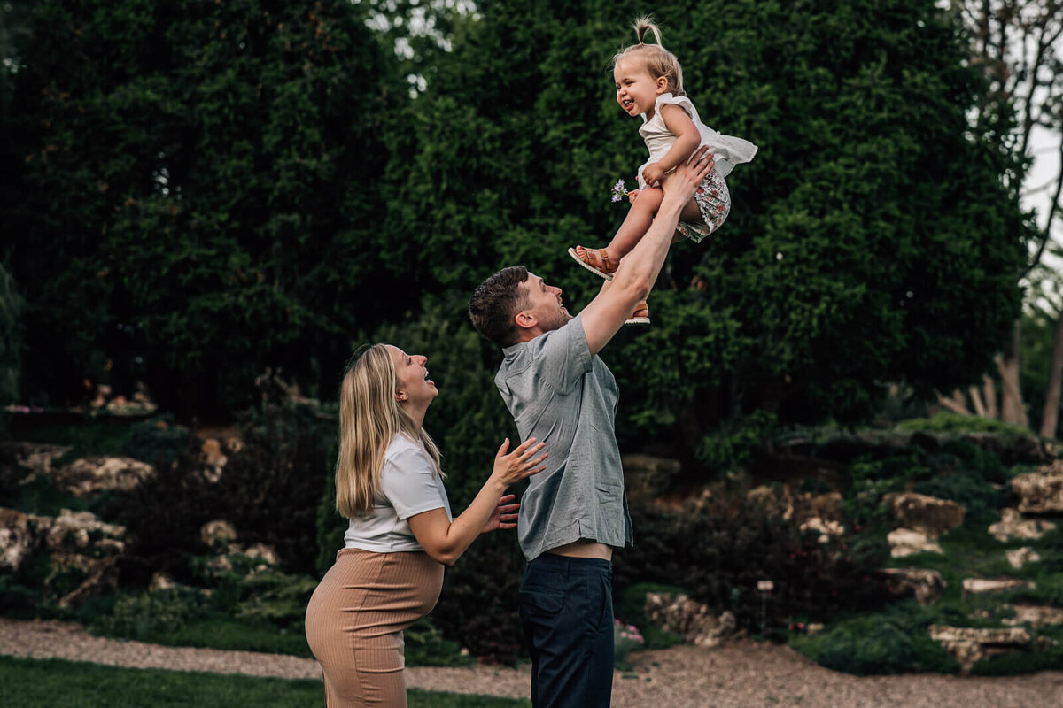 A father holds his laughing toddler high up in the air, while his expecting wife stands behind him reaching up and laughing.