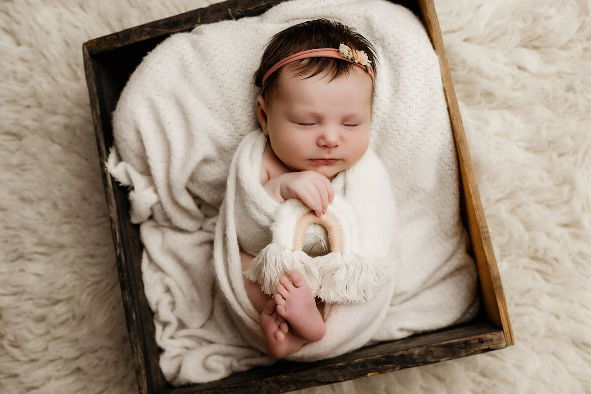 A swaddled newborn baby sleeps peacefully in a wooden box lined with white blankets, gently holding a handmade rainbow prop during a minimalist studio session in Scottsdale, Arizona.