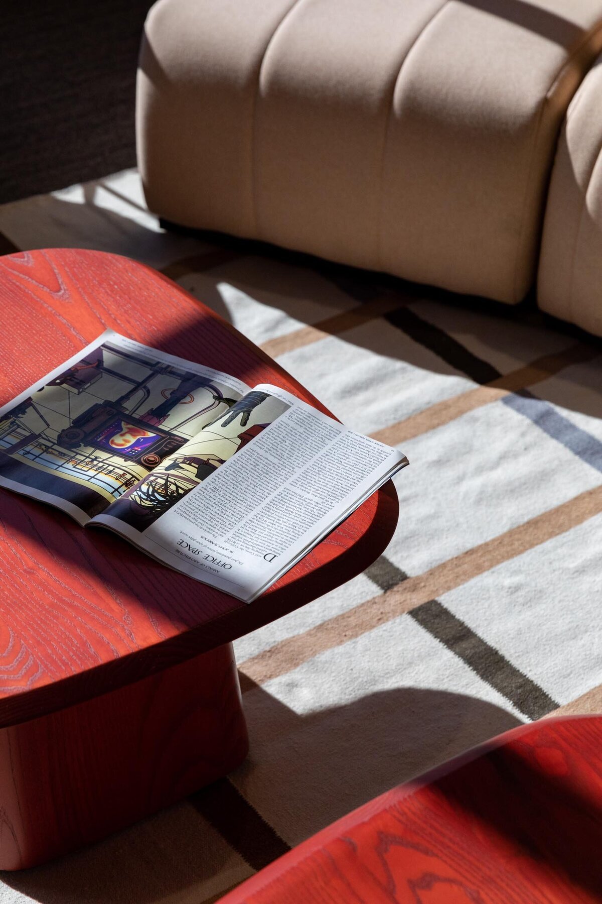 Magazine open on a red wood coffee table in a lounge area with a beige upholstered sofa and a geometric rug.