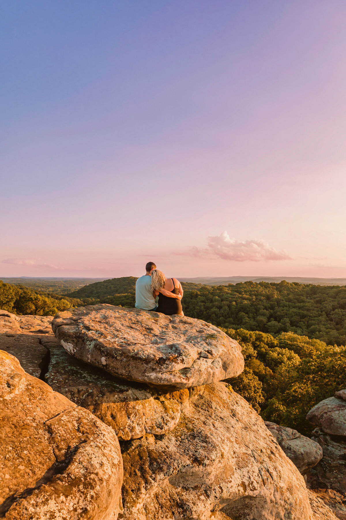 couple standing on a rocky overlook at Garden of the Gods in Southern Illinois during their engagement session, bathed in golden hour light with a panoramic view of the forest below. Engagement session 