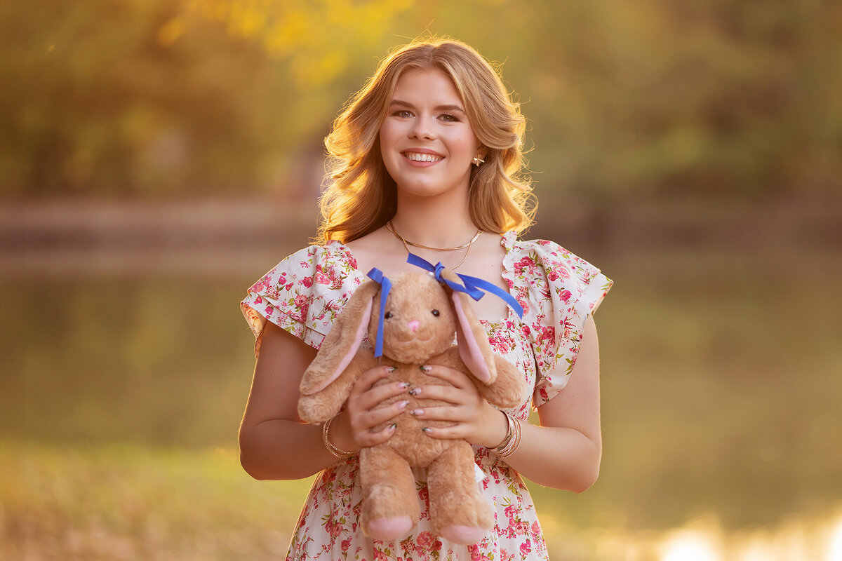 Dallas senior photographer captures a young woman in a floral dress, holding a stuffed bunny during golden hour by a peaceful river.