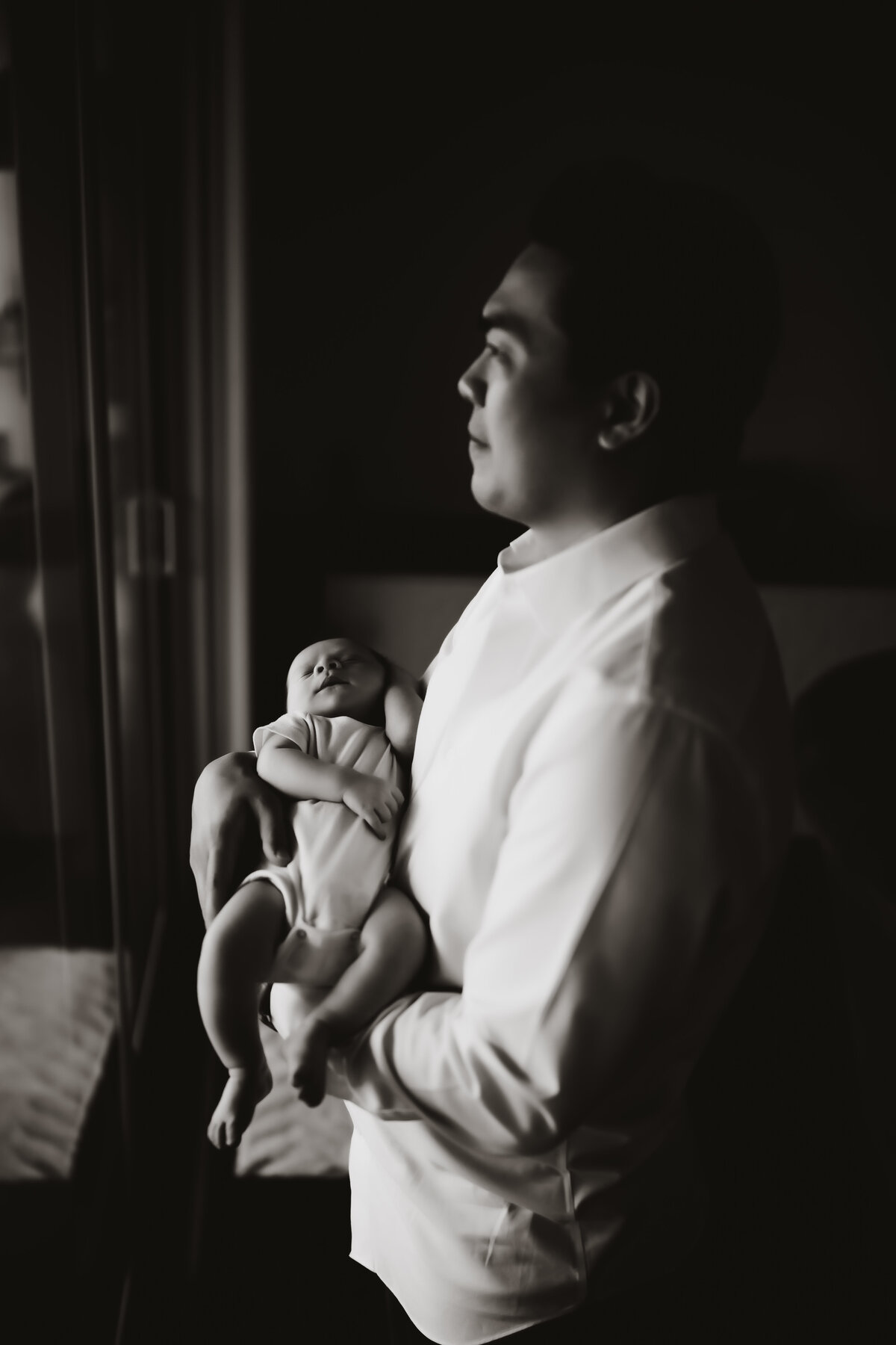 a black and white artistic portrait of a father holding his newborn baby in front of a large window