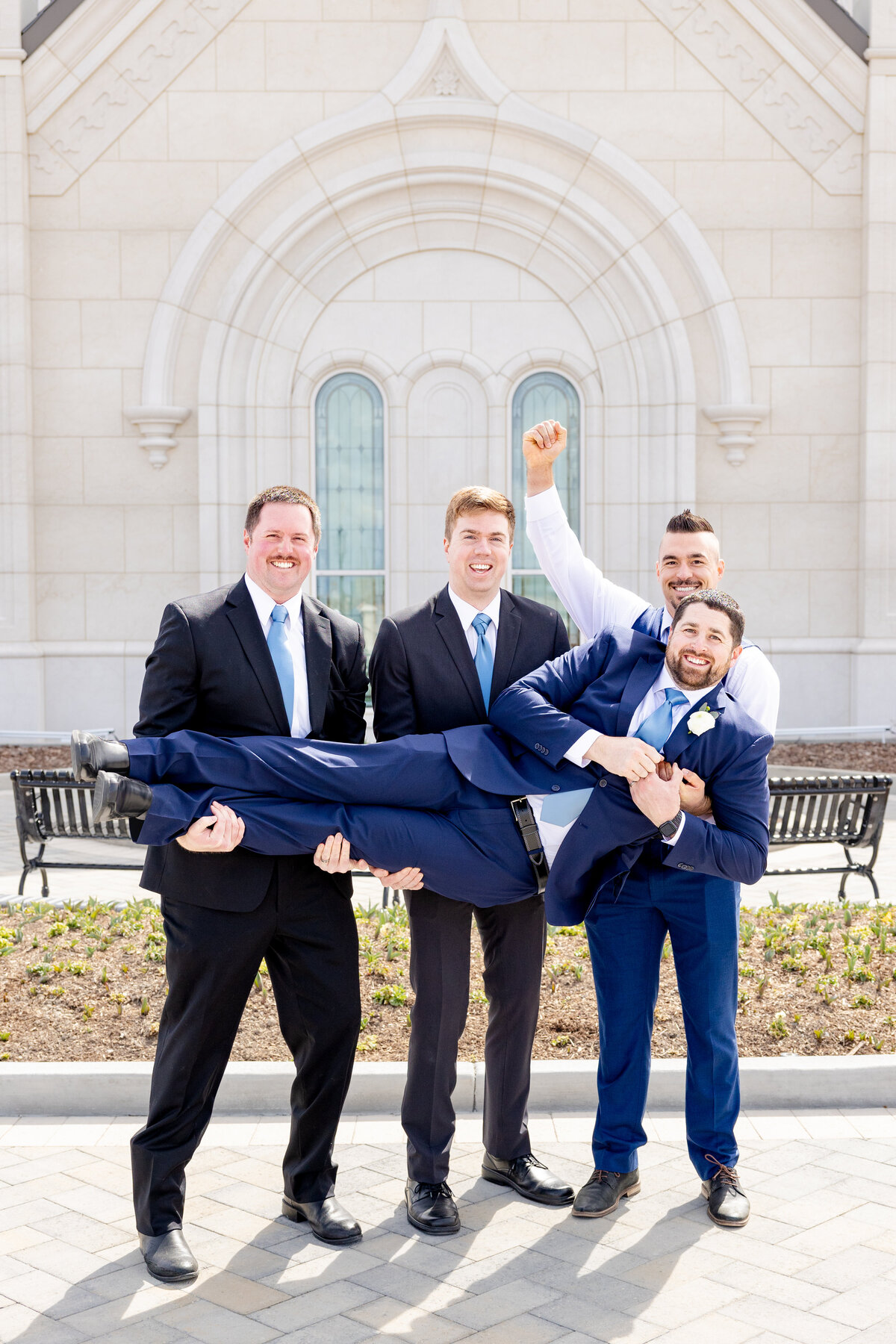 A fun moment where the groomsmen lift the groom off the ground, all laughing and celebrating outside the LDS temple.