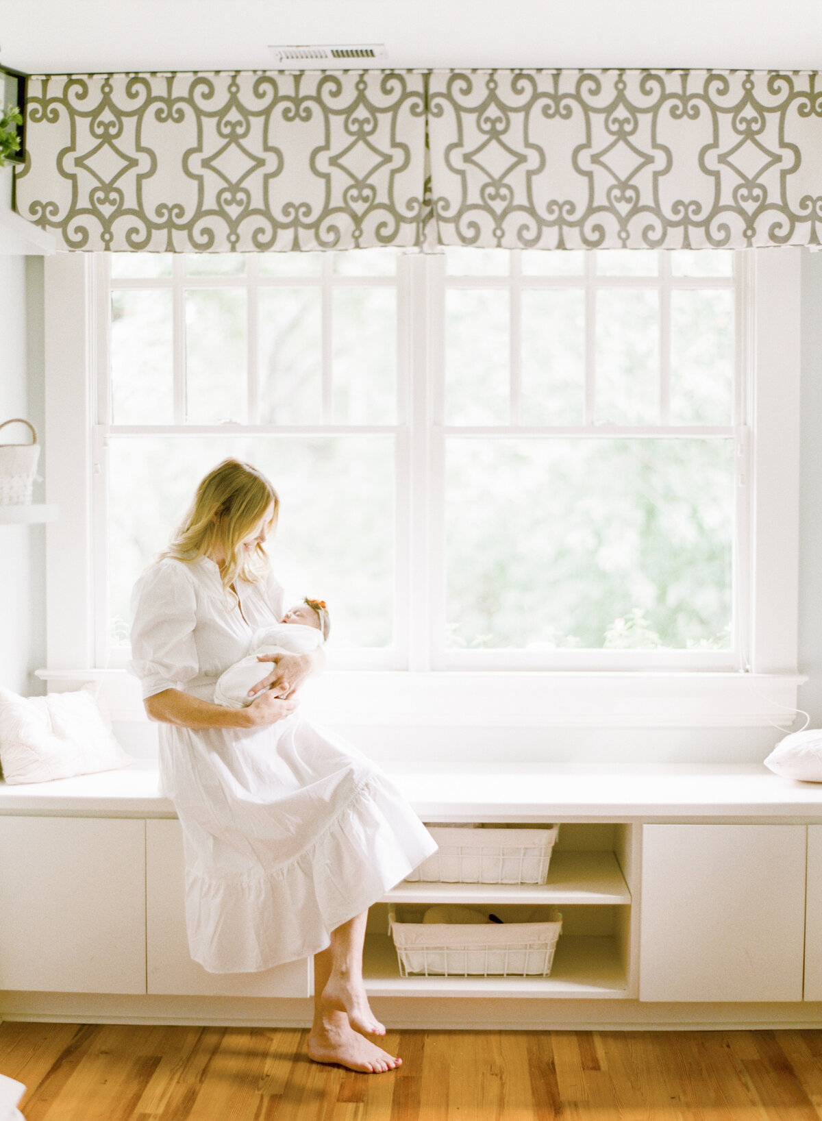 Mom sits in a large window seat and holds her newborn daughter. Image by Raleigh newborn photographer A.J. Dunlap Photography.