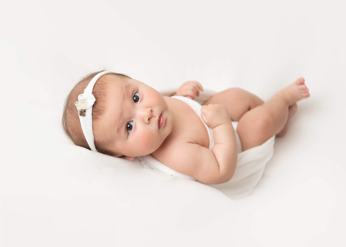 3 month old baby girl laying on a white backdrop in a white swaddle looking back at the camera - By Los Angeles Newborn Photographer