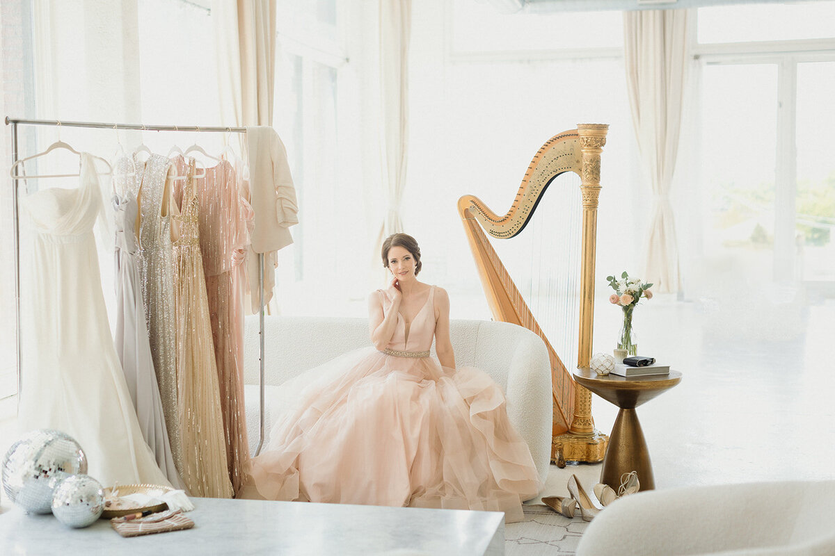 woman in light pink ballgown is stilling on a couch with a harp behind her. There is a rack of gowns next to her and she is looking at camera and smiling