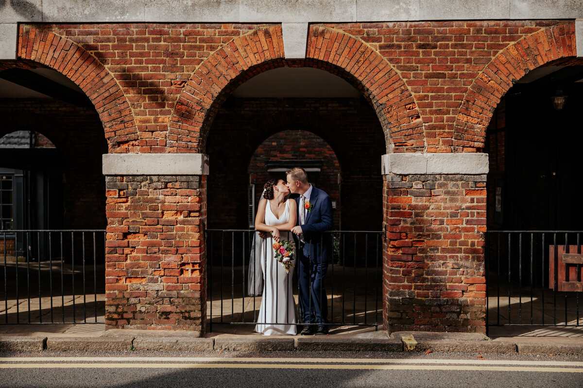 Bride and groom under an archway, kissing while leaning against a railing in Old Amersham high street