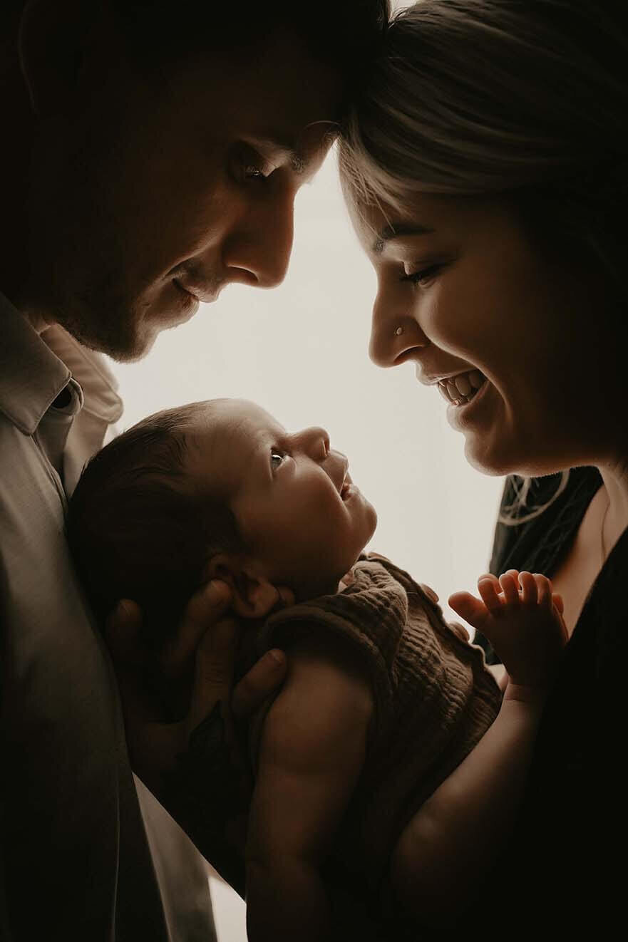 a portrait of a family of three where mom and dad look down at their new son who smiles up at them