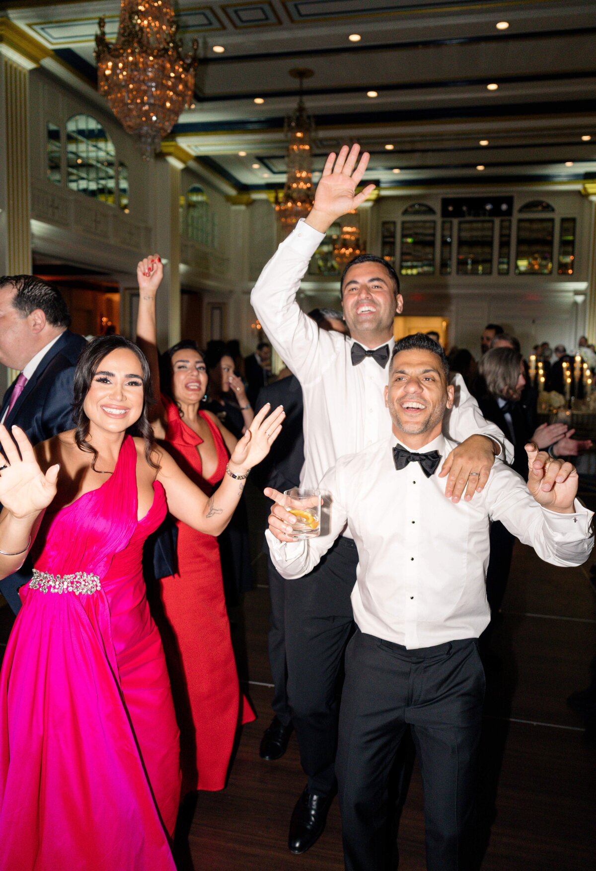 A group of people, dressed in formal attire, are joyfully dancing at a lively event in an elegant venue. A man in a white shirt holds a drink. The atmosphere is festive, with chandeliers and large windows visible in the background.