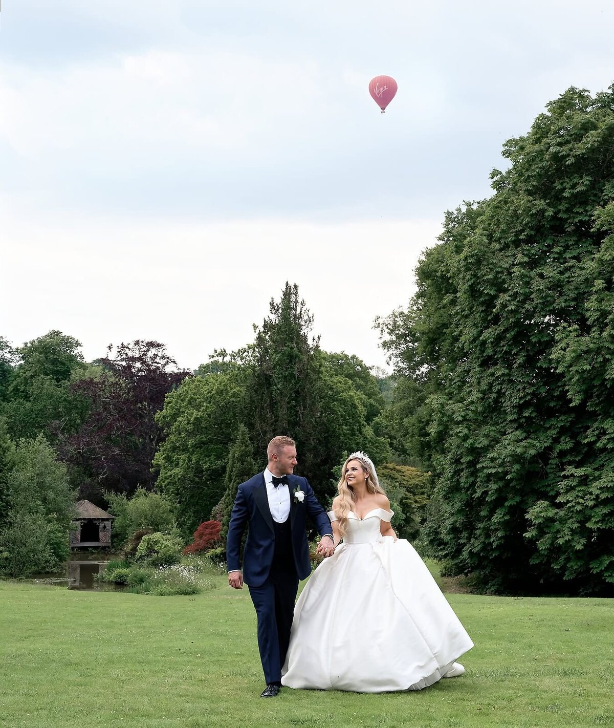 Bride and groom walking and smiling in the grounds of The Ravenswood with a hot air balloon in the sky above them