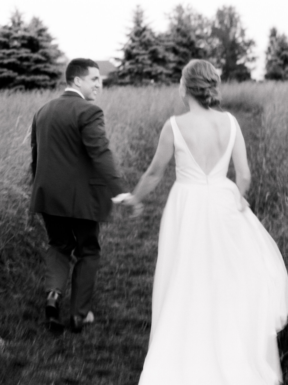 Black and white photo of bride and groom holding hands and walking through a grassy field
