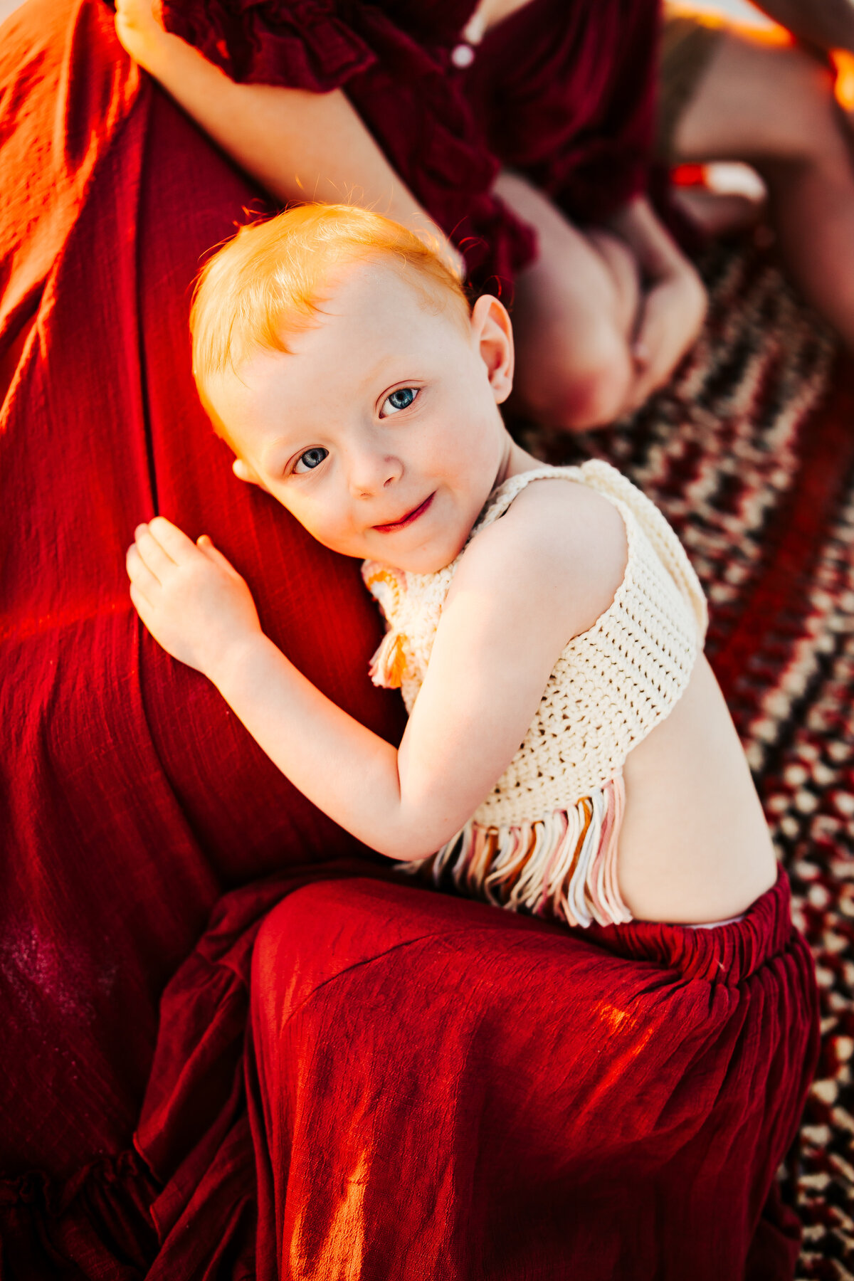 a red headed toddler wearing a boho outfit laying on her mom at the beach