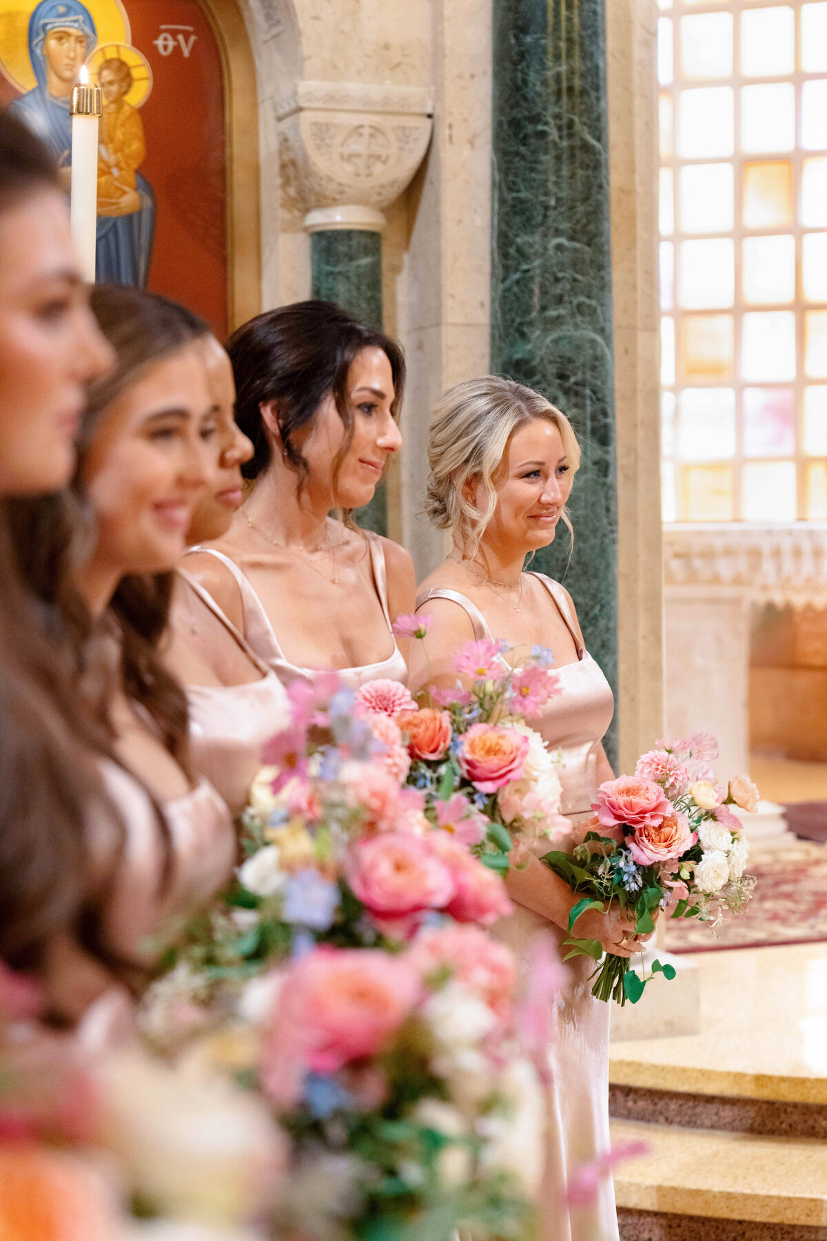 A group of women in elegant dresses stand holding colorful bouquets. They are indoors, possibly in a church, suggested by the decorative stone pillars and a religious icon visible in the background. The atmosphere is formal and celebratory.