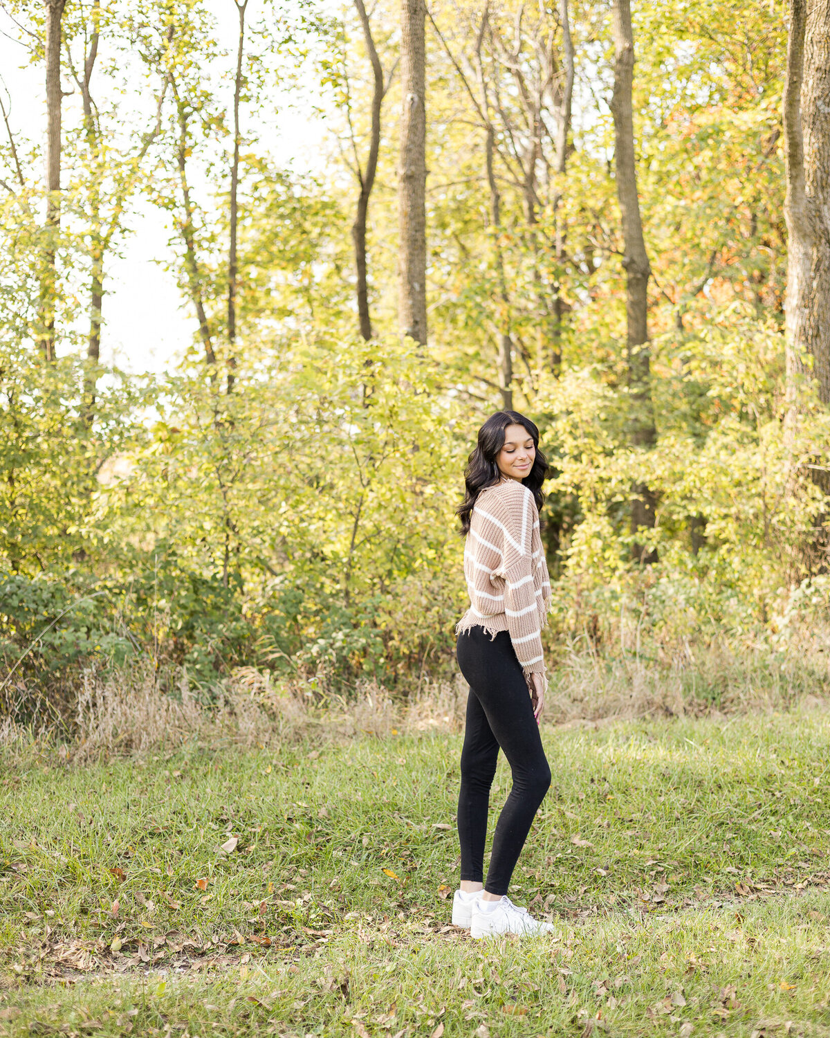 high school female senior, with long black hair, standing in grass with a tree line next to her, standing to the side, looking down smiling softly, wearing black leggings, and light tan and white stripped shirt