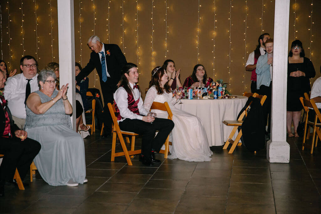 A bride and groom sit with guests at round tables, enjoying a wedding reception. String lights hang in the background, creating a warm, festive atmosphere. Some guests are smiling and clapping.