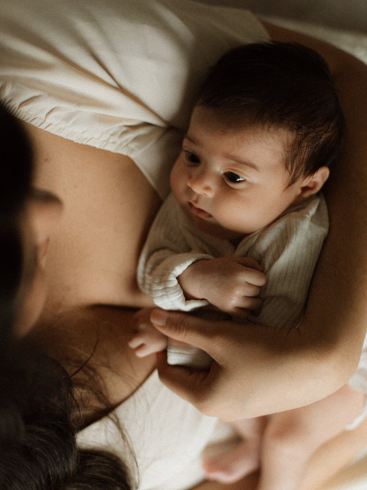 Mother holding newborn by a bright window in an intimate in-home session with soft light and neutral tones.