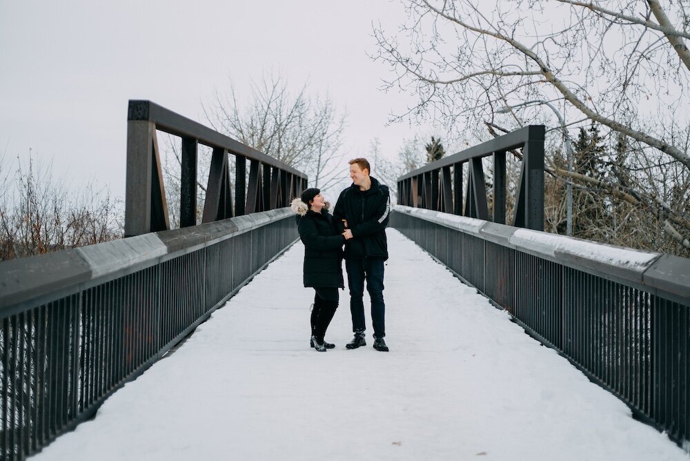 An man and a woman embrace on a bridge covered in snow in their black coats