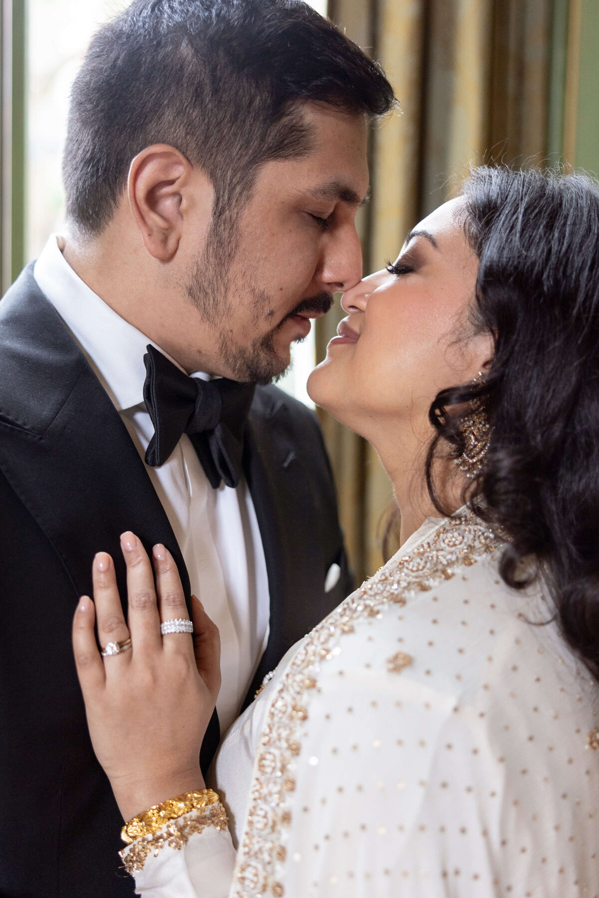 A couple dressed in elegant attire shares an intimate moment, standing close with their foreheads nearly touching. The man wears a black tuxedo, and the woman is in a white outfit with gold accents, smiling with her hand on his chest.