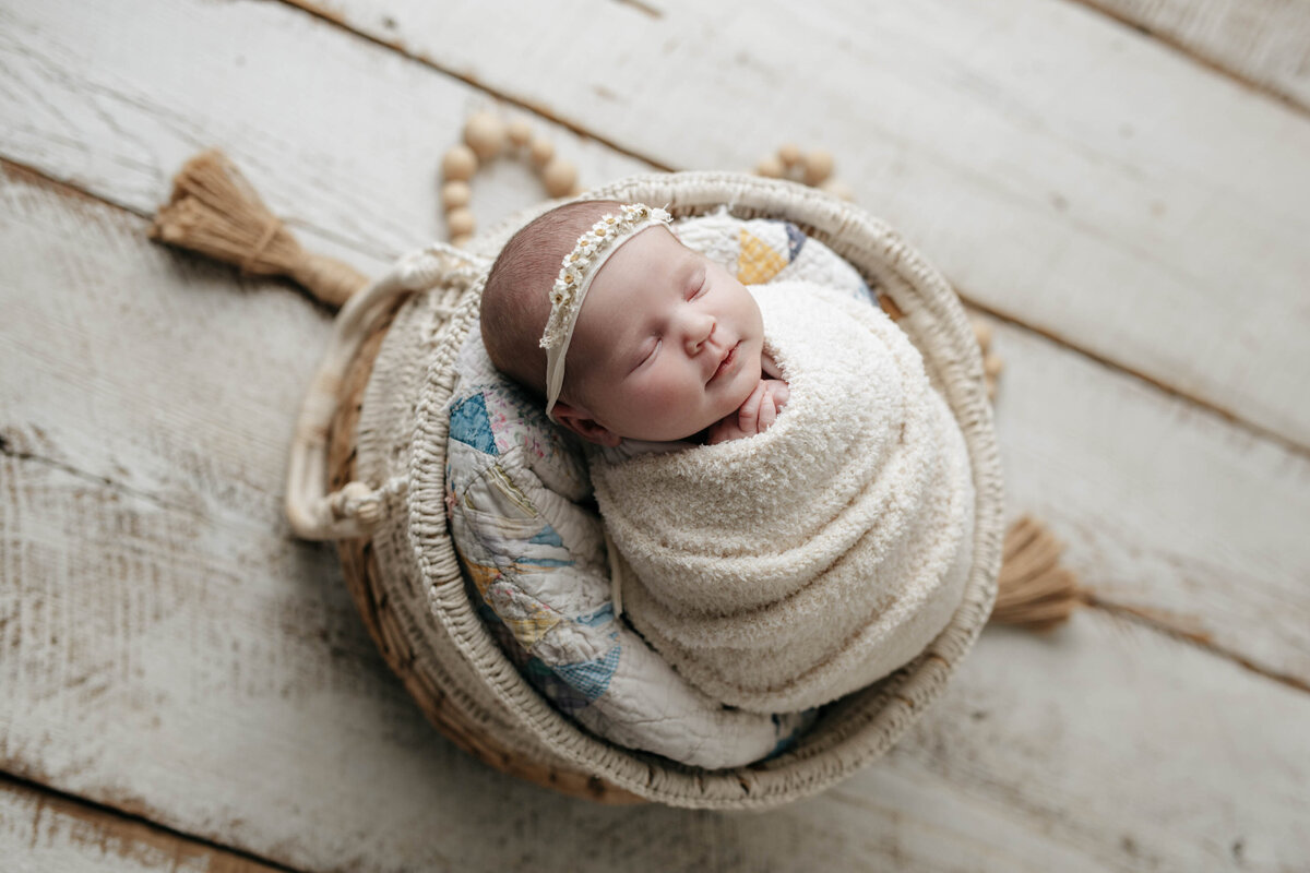 Studio newborn photography | Sleeping baby swaddled in cream wrap in basket. Baby wearing a delicate cream headband with hands clasped under chin.