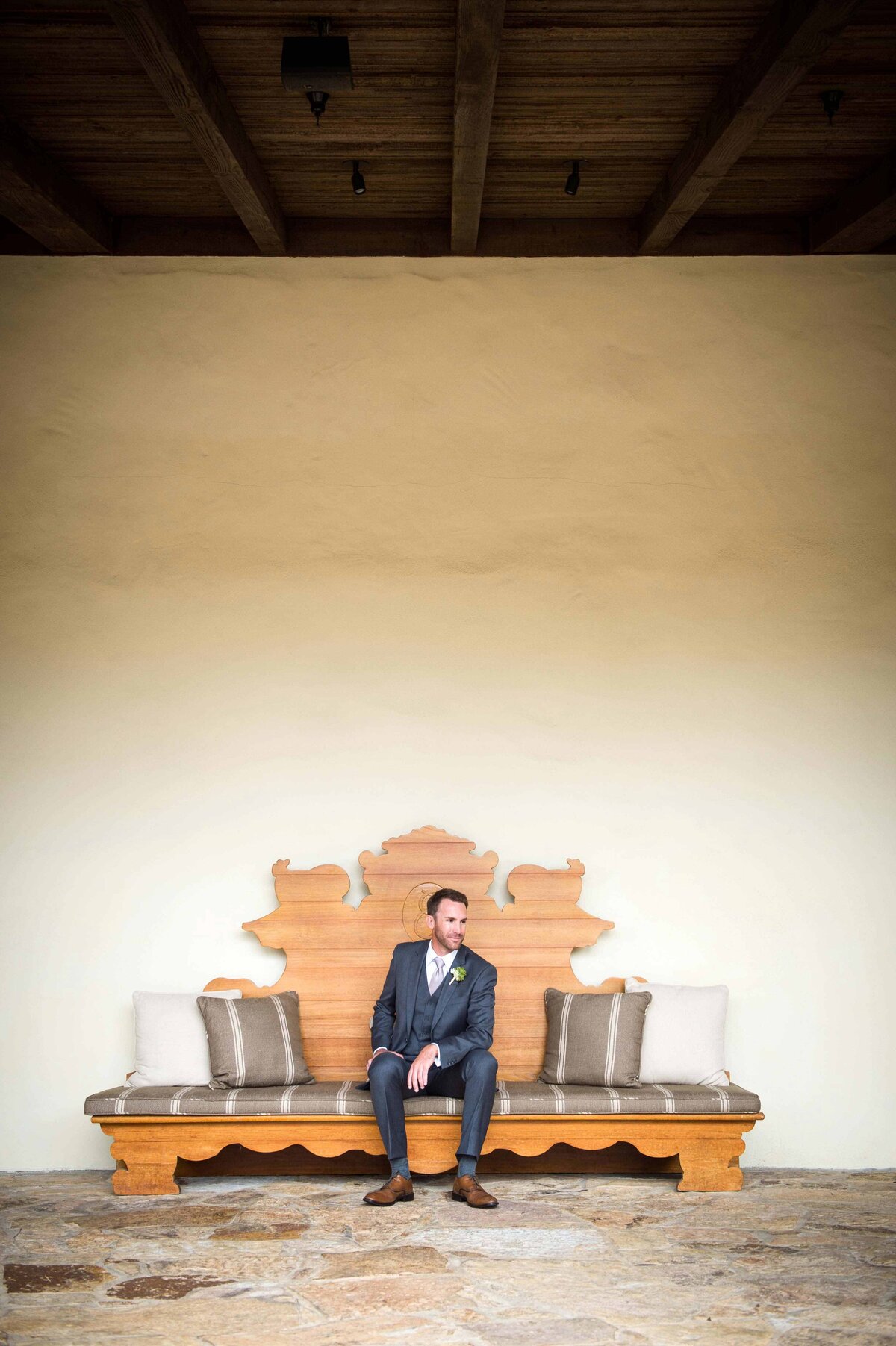 A man wears a formal tailored suit while seated on a wooden bench at the Estancia La Jolla Hotel