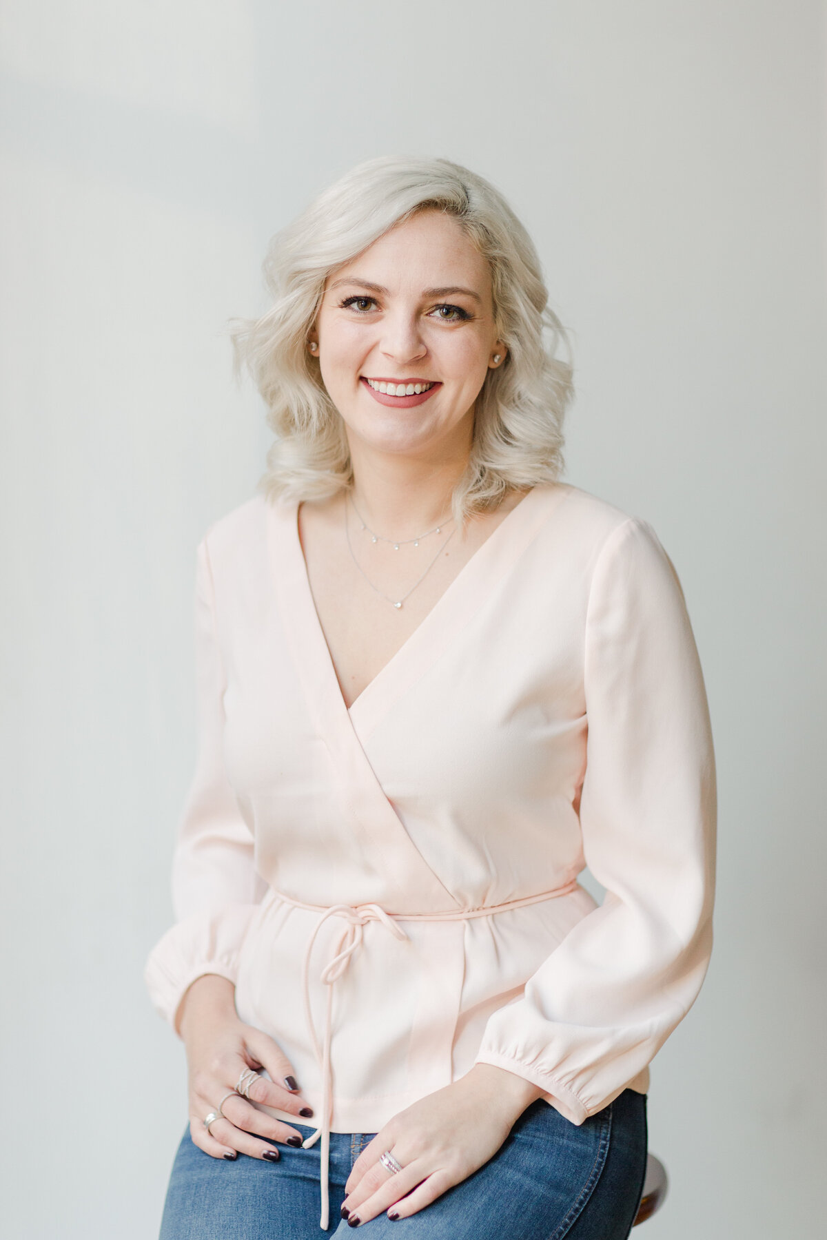 blong woman in pink top looking at camera and smiling as she sits on a stool