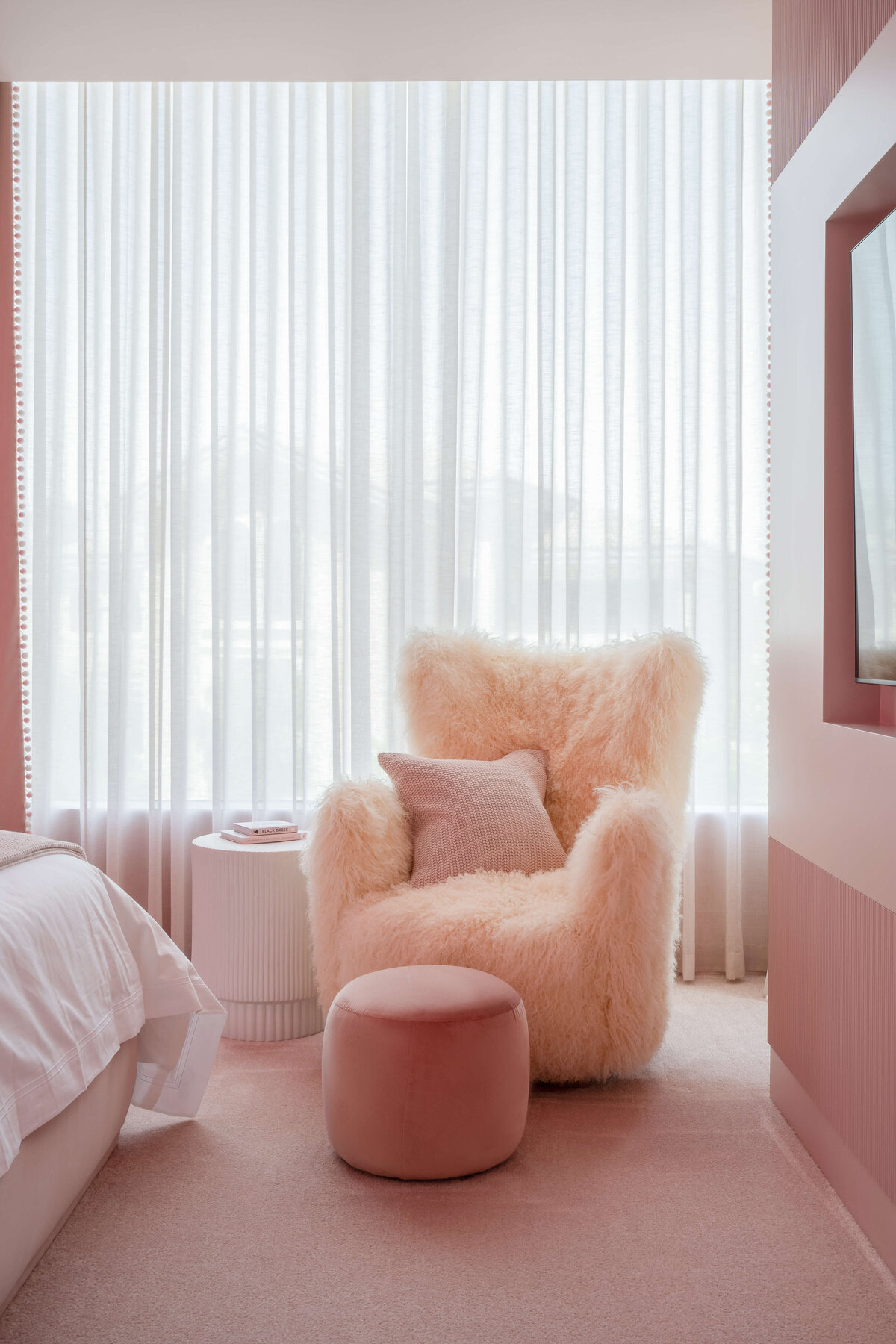 Soft pink furry chair in girl's bedroom with a darker pink stool in front. Behind the chair is a large window covered with sheer white curtains.