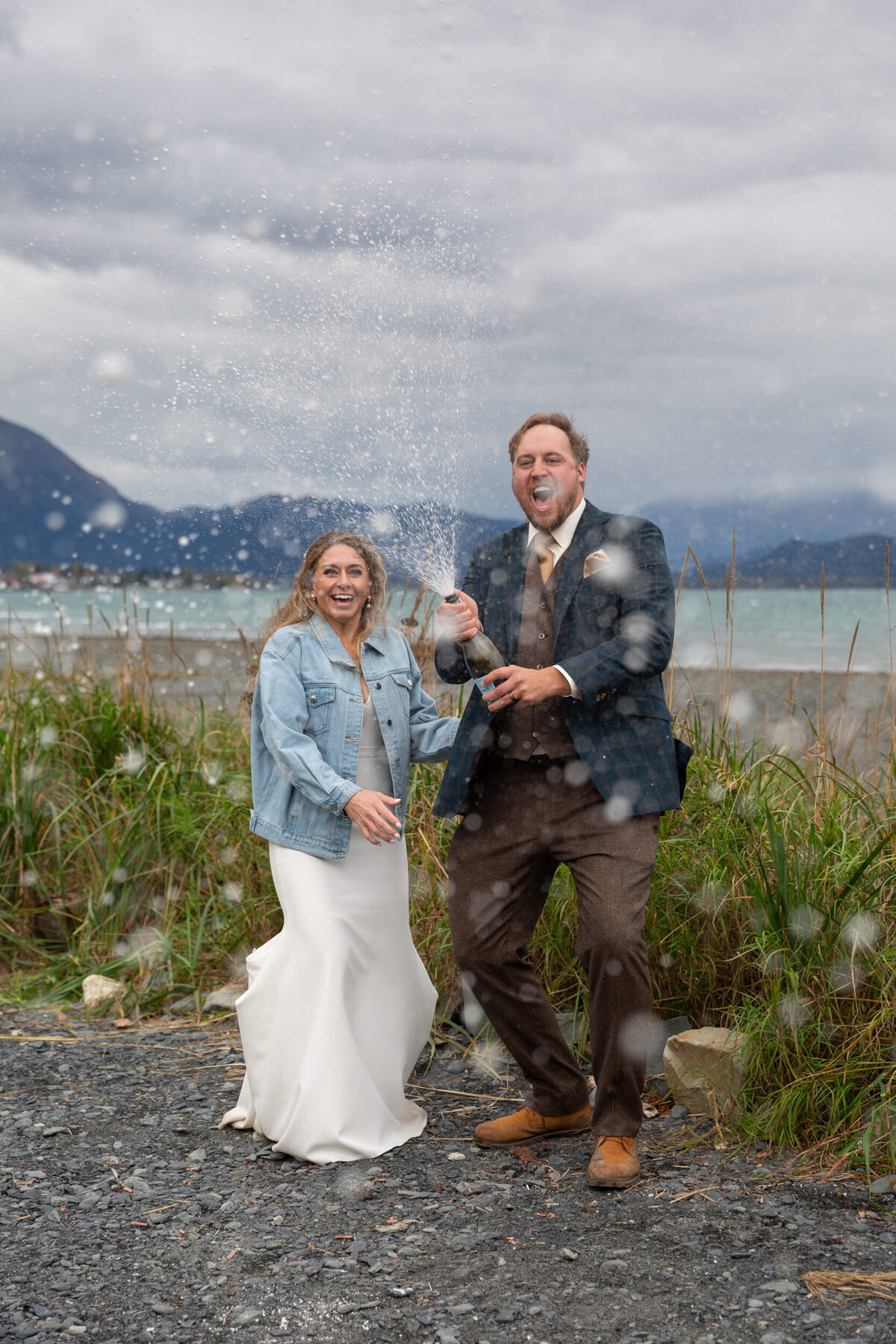 A bride and groom spray a bottle of champagne directly at their Alaska elopement photographer.