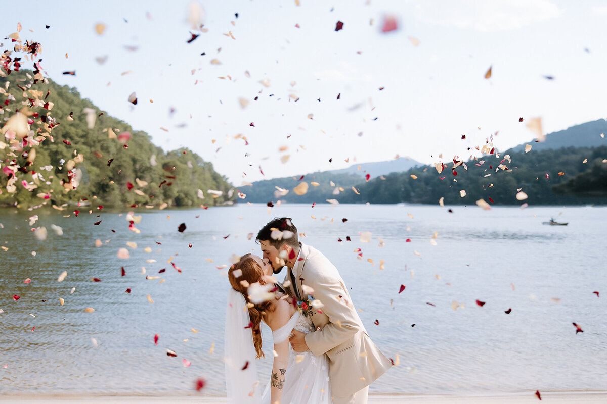 Bride and Groom kiss lakeside as flower pedals are thrown around them