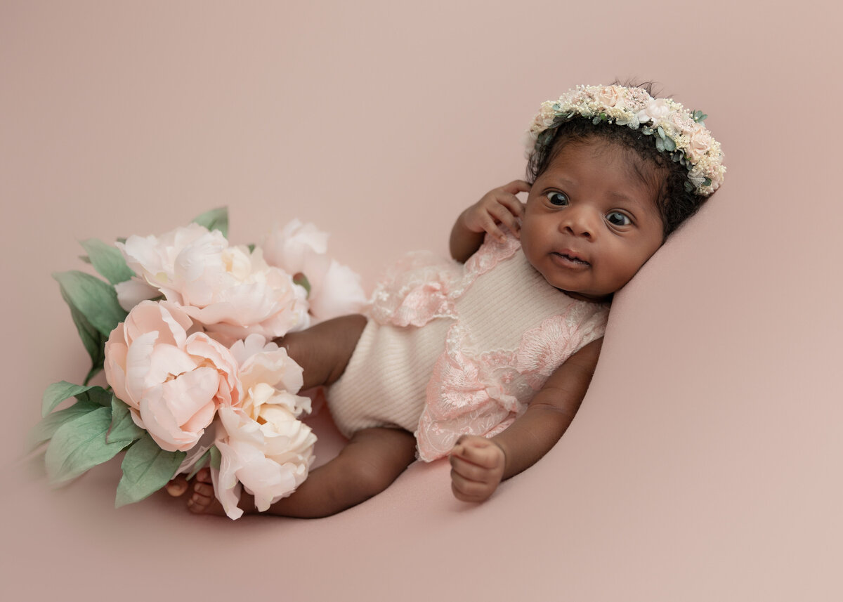 A newborn baby with eyes open lays in a pink oneie and white floral headband in a studio