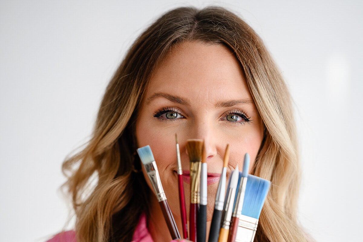 A brand photographer in Denver takes a photo of an artist holding up paint brushes in front of her face during her branding photos during her branding photo shoot.