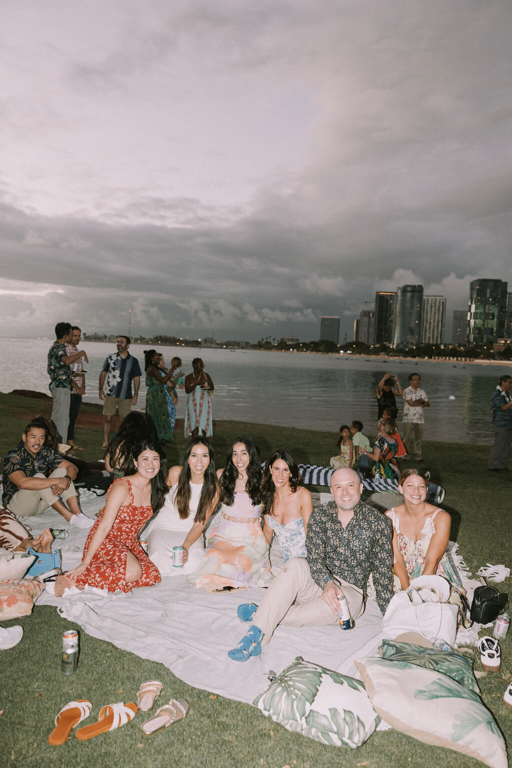 Nighttime shot of wedding guests at welcome dinner smiling in front of the ocean and honolulu cityscape