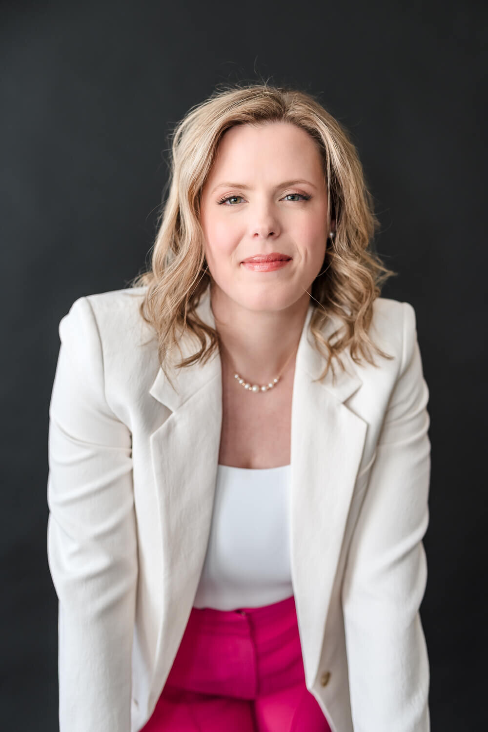 Poised female entrepreneur leaning forward slightly, wearing white blazer and pink trousers in Kelowna photo studio.