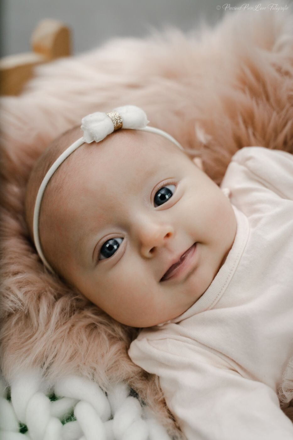 Close-up of a baby wearing a pink knitted hat with a red flower, gazing curiously with wide dark eyes.
