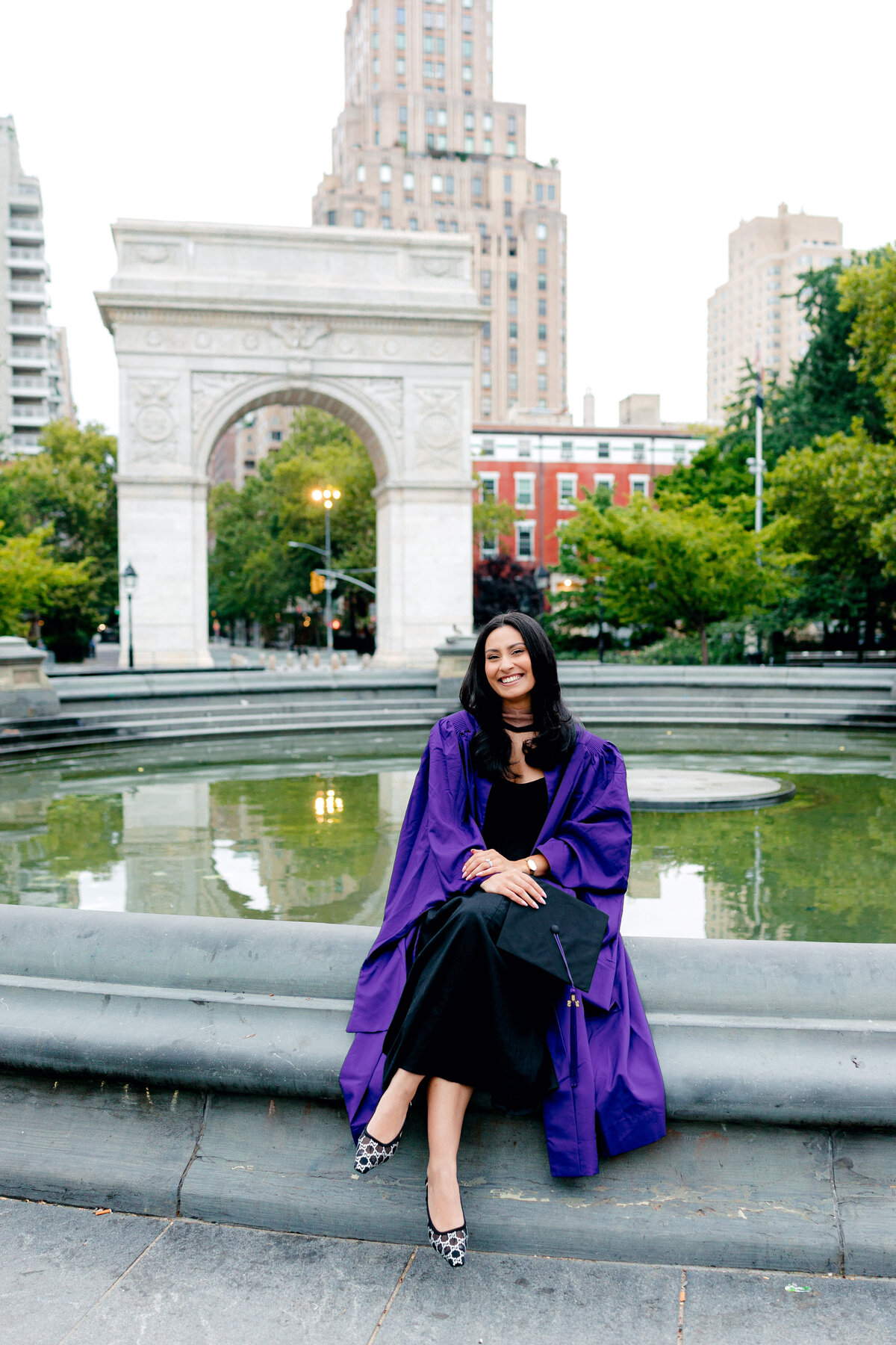 NYU graduate smiling in purple gown and cap while sitting by the fountain at Washington Square Park.