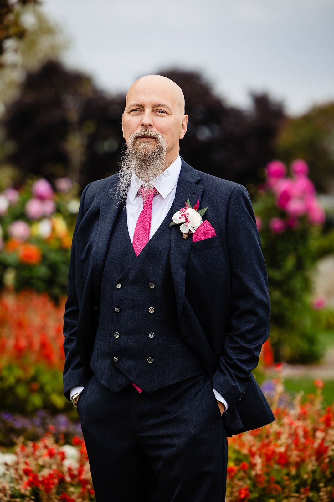 A groom standing confidently in a formal blue suit with a pink tie and a flower boutonnière, posing in front of a vibrant flower garden