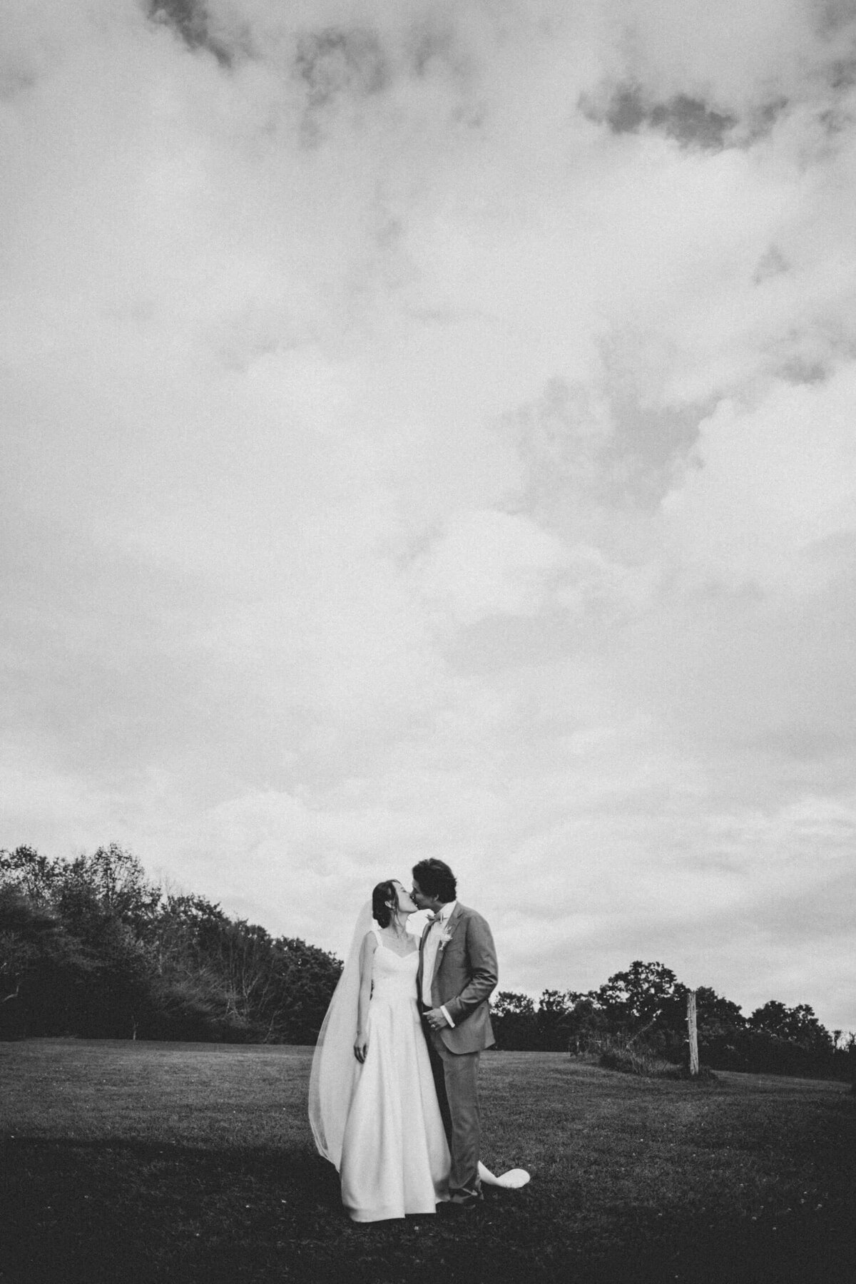 A black-and-white portrait of a bride and groom sharing a kiss under a vast cloudy sky