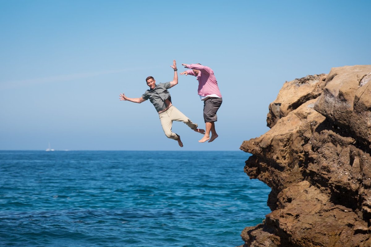 Two brothers jump off a large rock and are mid-air heading towards the ocean waters