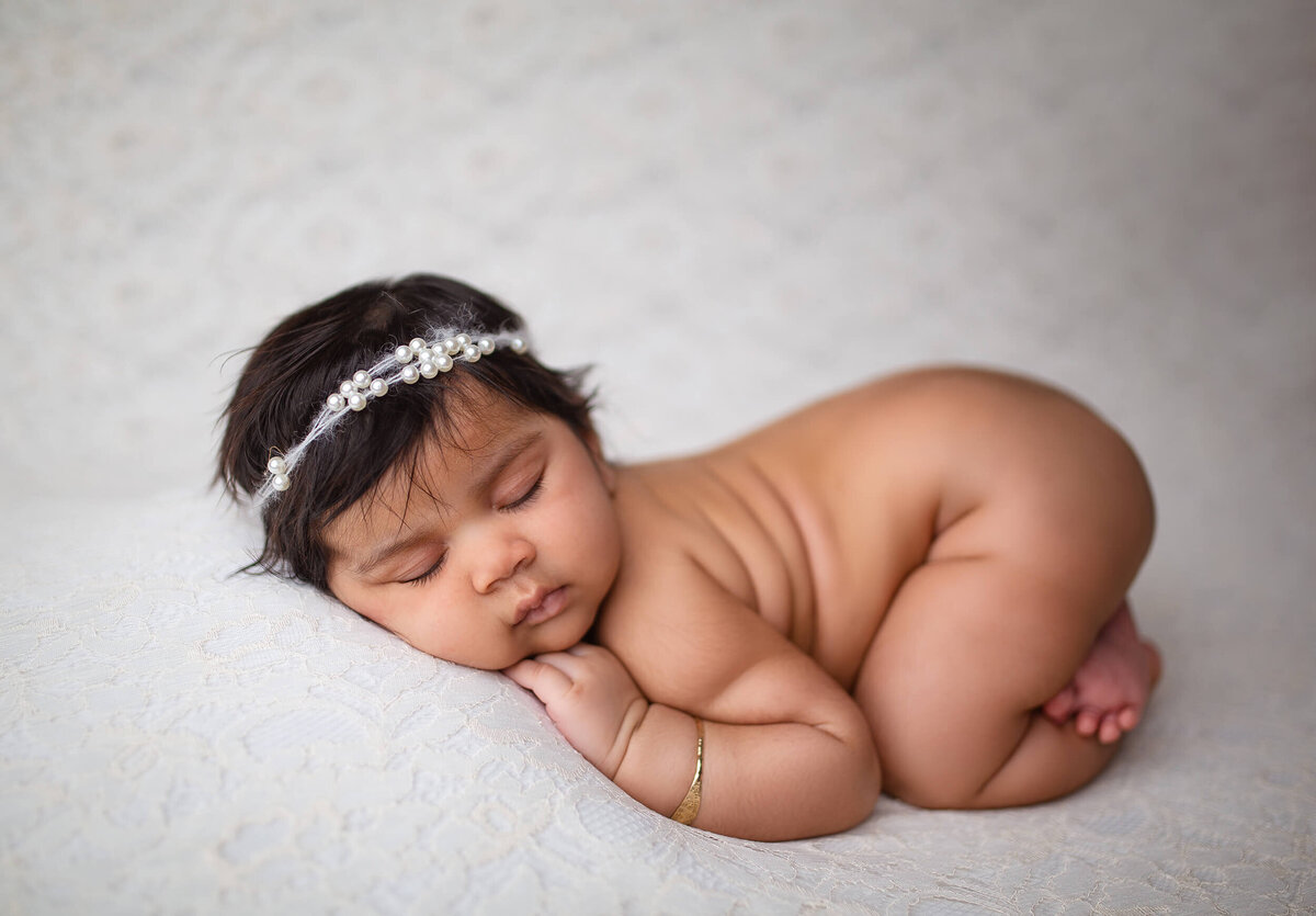Beautiful brown haired chubby sleeping baby in studio posed on white lace with pearl headband. Captured by newborn photographer Sarah Wyndham Photography