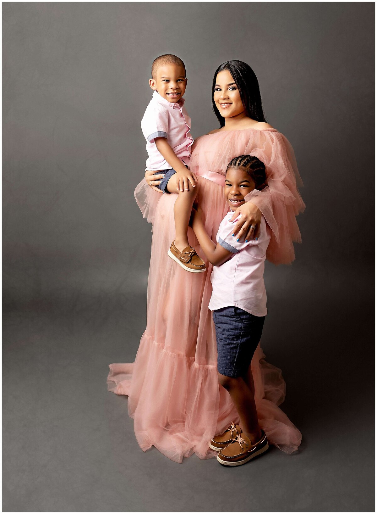 A mother, heavily pregnant stands holding her younger son while the other son stands next to her in a maternity portrait. The older boy lovingly embraces his mother's baby bump, creating a heartwarming and joyful family moment.