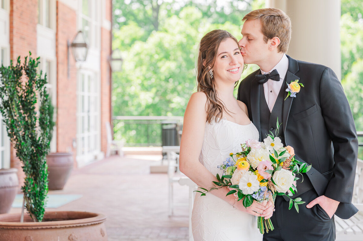 A loving groom giving his new bride sweet kisses on her temple on a beautiful brick porch at The Prestonwood Country Club in Cary as they enjoy their Raleigh wedding photography session by JoLynn Photography